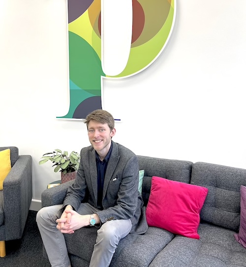 A young man sat on a sofa with a large P logo in the background on a white wall