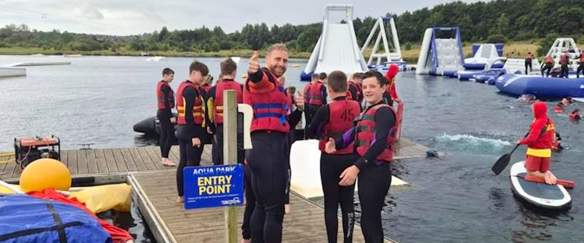 A group of young people stand on a jetty lakeside in wetsuits ready to start an activity