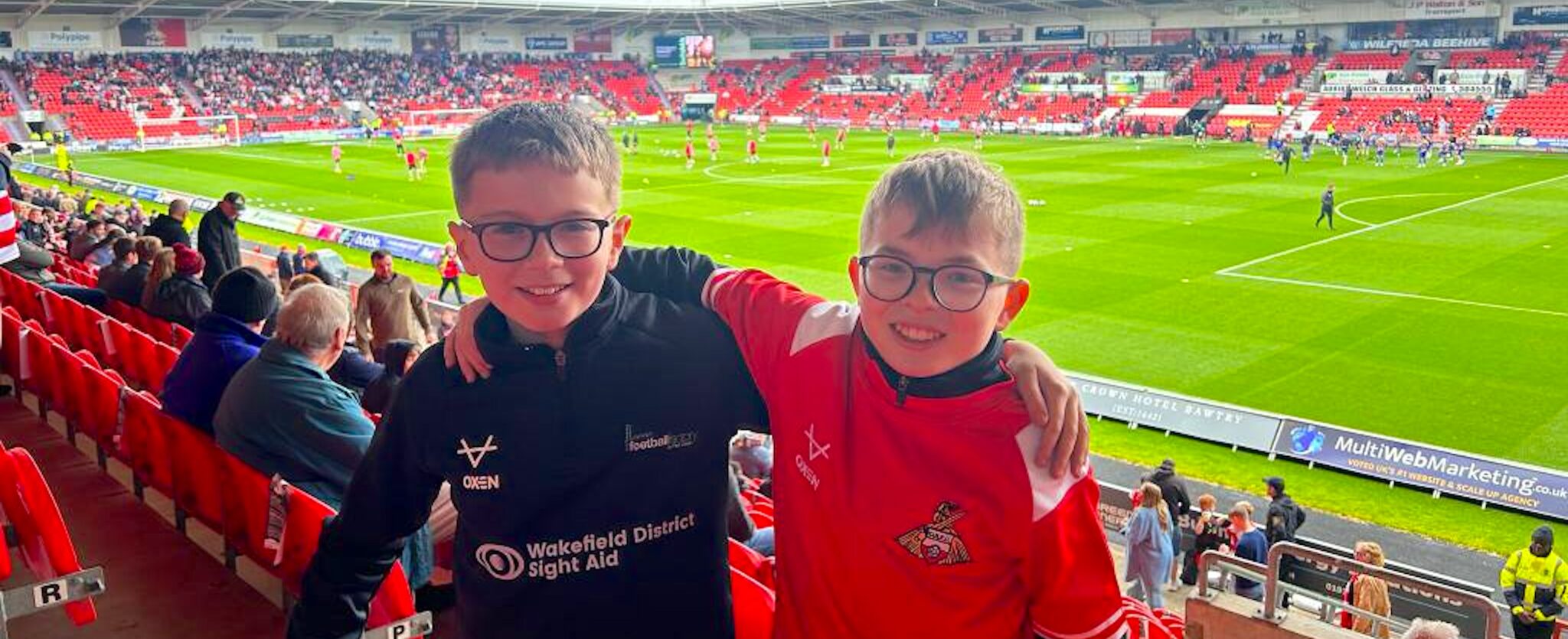 Two young boys stand arm in arm at a football match with smiles, they wear glasses and sports tops