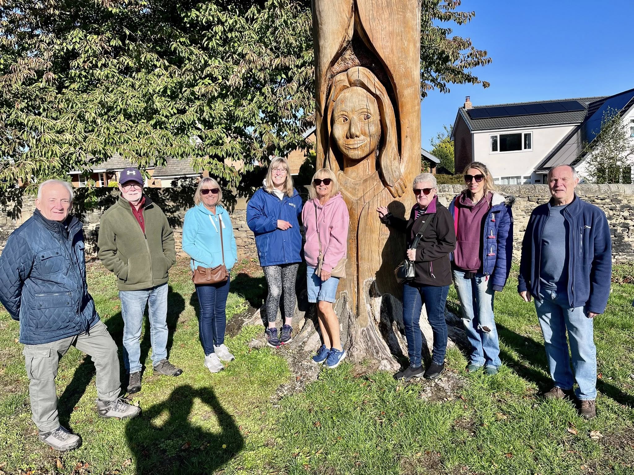 A group of mixed aged people stand next to a wooden carving in Carr Lodge Park, Horbury