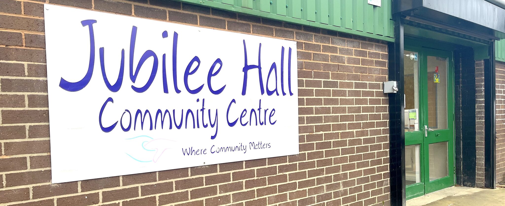 The outside of a community centre with a sign stating 'Jubilee Hall' leading to two green entrance doors