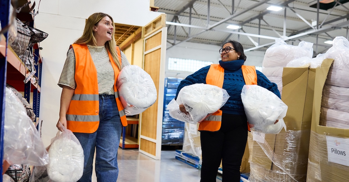 Two women wearing high viz walk through a warehouse carrying bedding and smiling