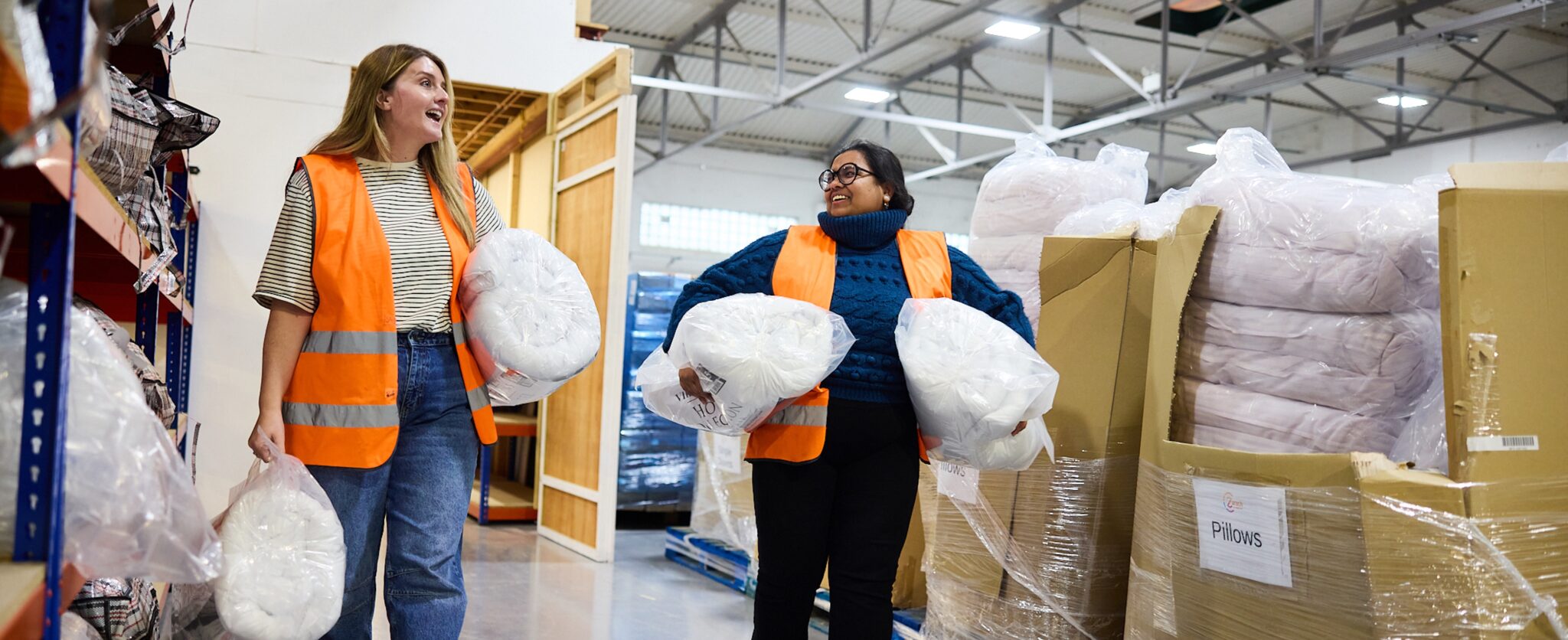 Two women wearing high viz walk through a warehouse carrying bedding and smiling