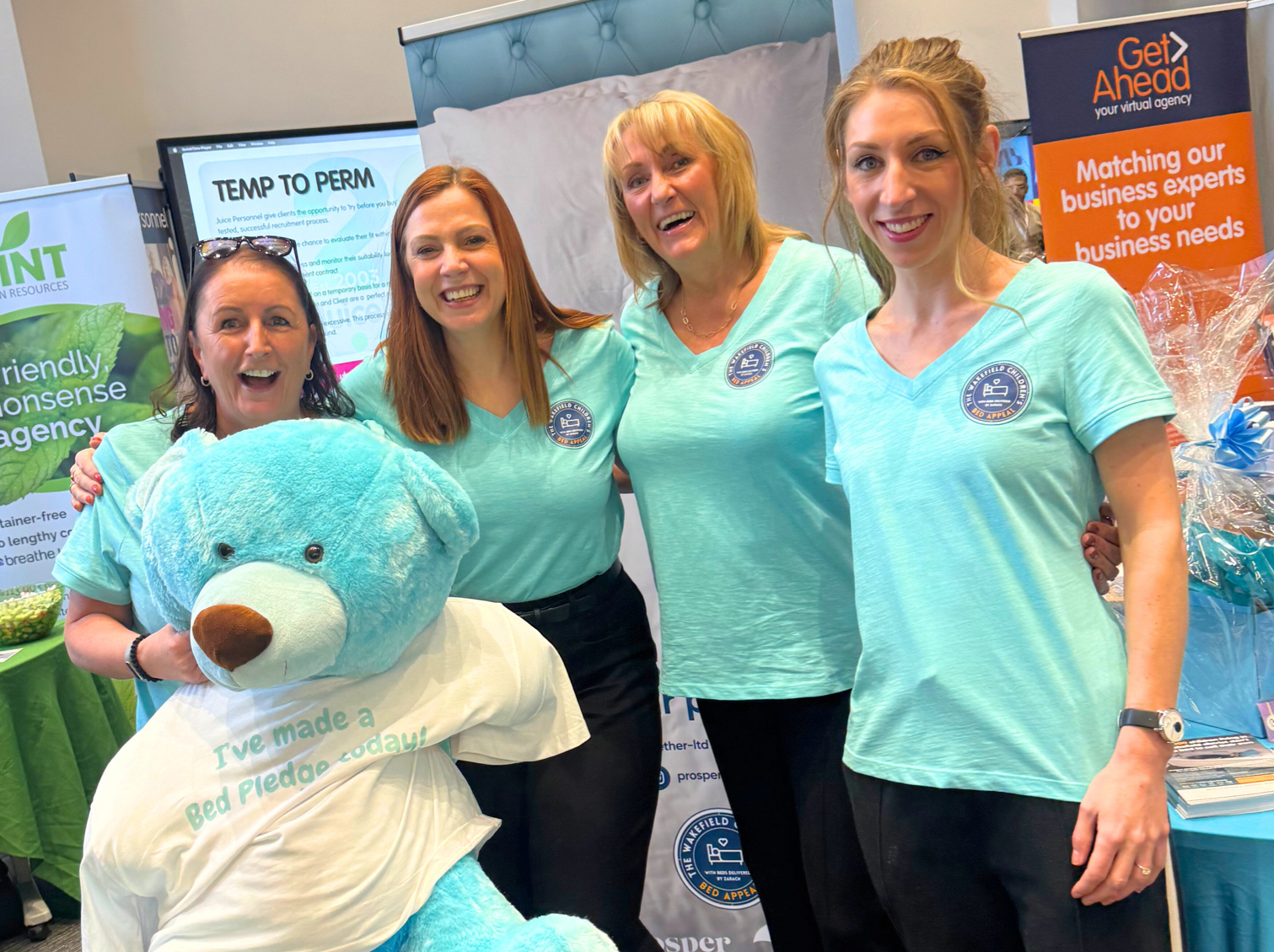 4 women wearing Bed Appeal t-shirts stand in front of an appeal banner raising money